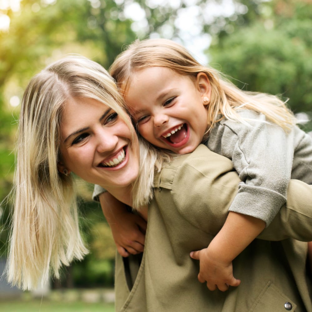 Mom and her daughter smiling while mom carries her daughter on her back after visit to Village Orthodontics